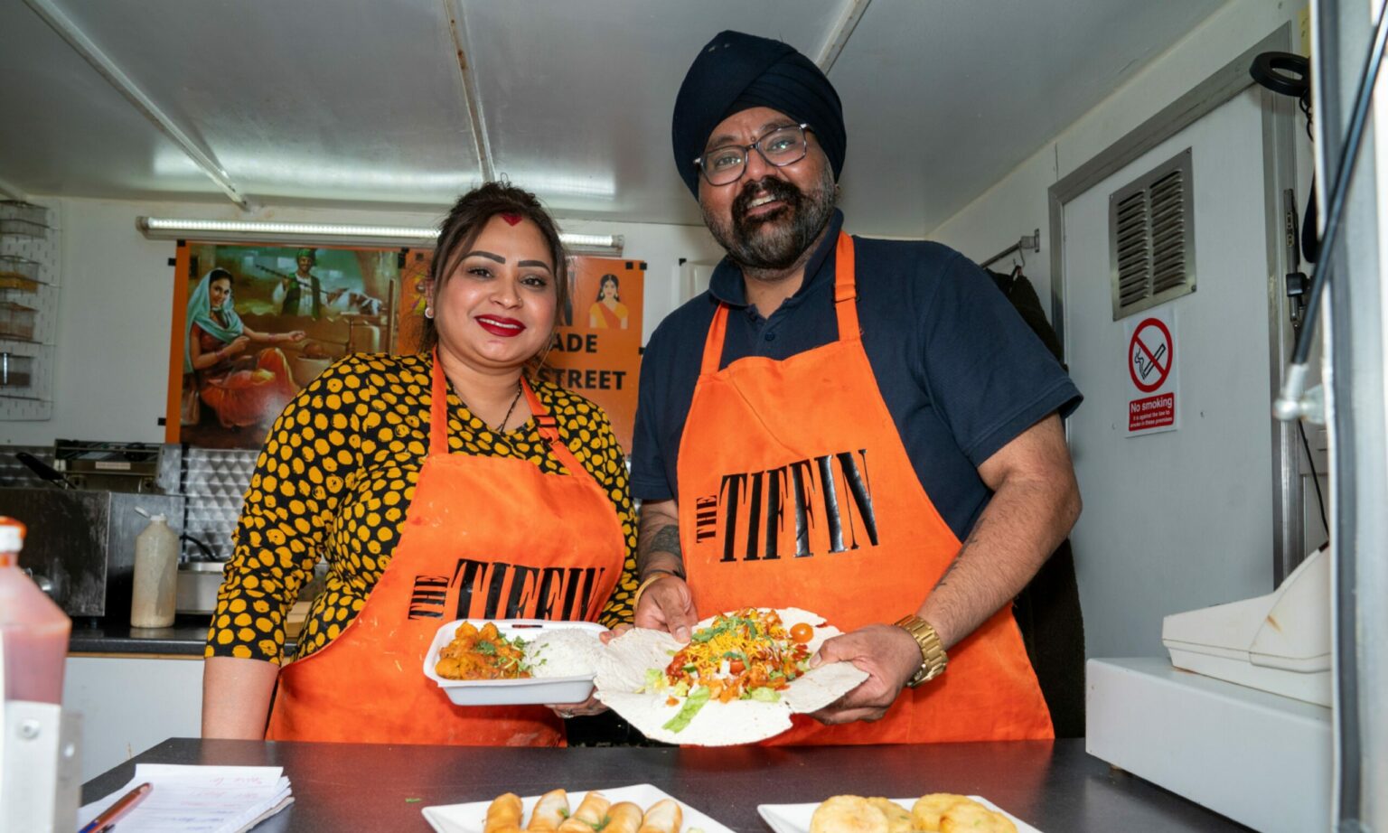 Tony and Priya in orange Tiffin aprons in a kitchen, smiling. Priya holds curry and rice, and Tony holds a large open flatbread with pakoras and salad.